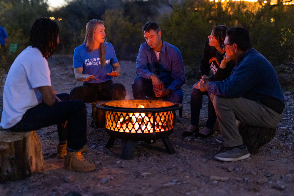 Five people on stumps around a fire pit at dusk, one of them speaking with her hands, the others listening