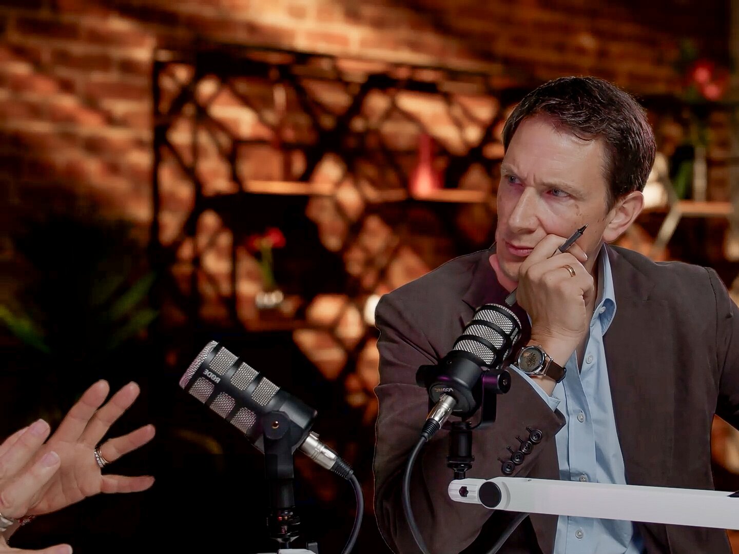 Chris Kutarna listening across a studio table, pen to his chin, as a guest gestures at the edge of the frame